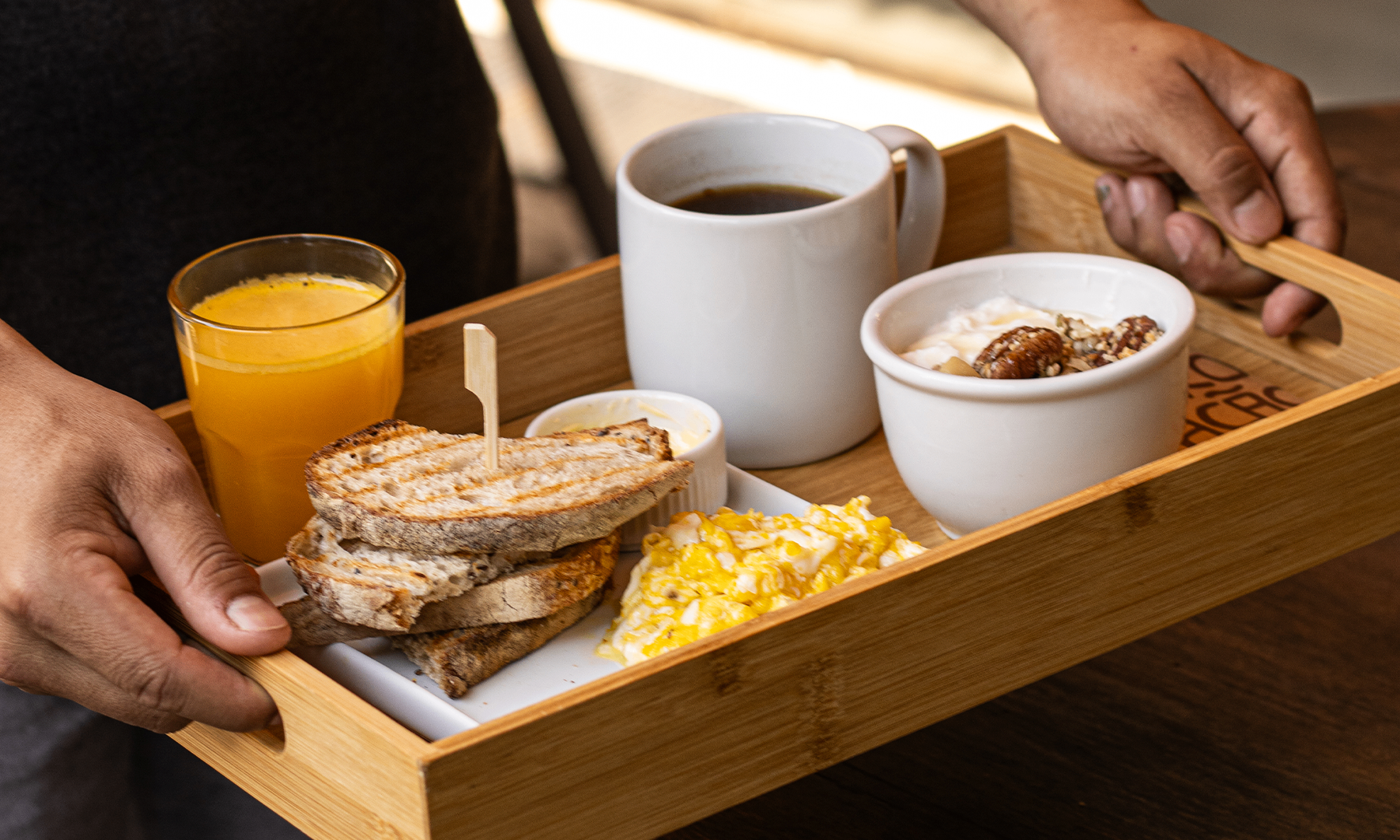 Bandeja de madeira com café preto, suco de laranja, ovos mexidos, torradas do pão da casa, manteiga e pote de iogurte com granola e castanhas, servida no Yarn.