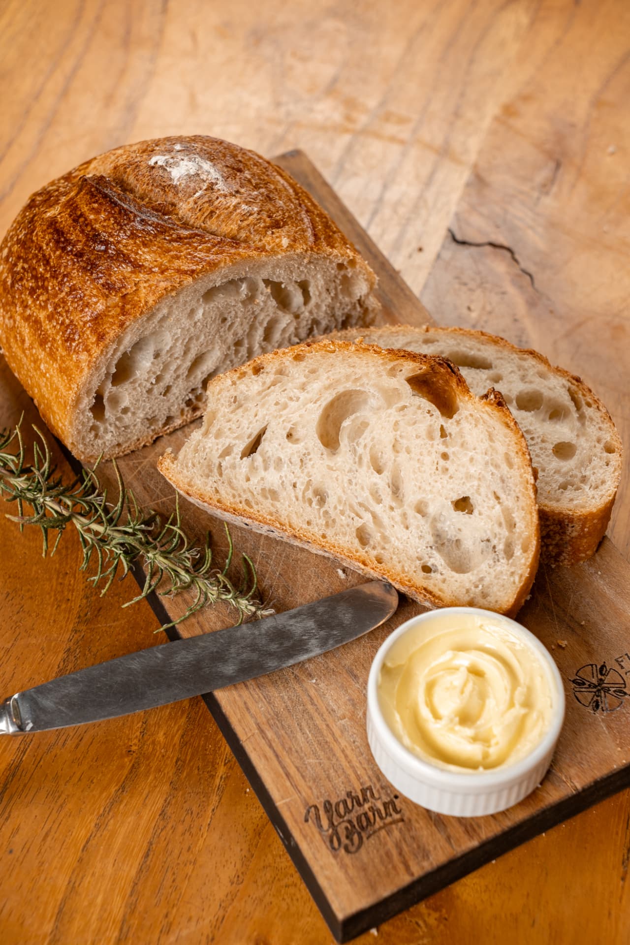 Pão de fermentação natural do Yarn cortado em fatias sobre tábua de madeira, com manteiga e alecrim.