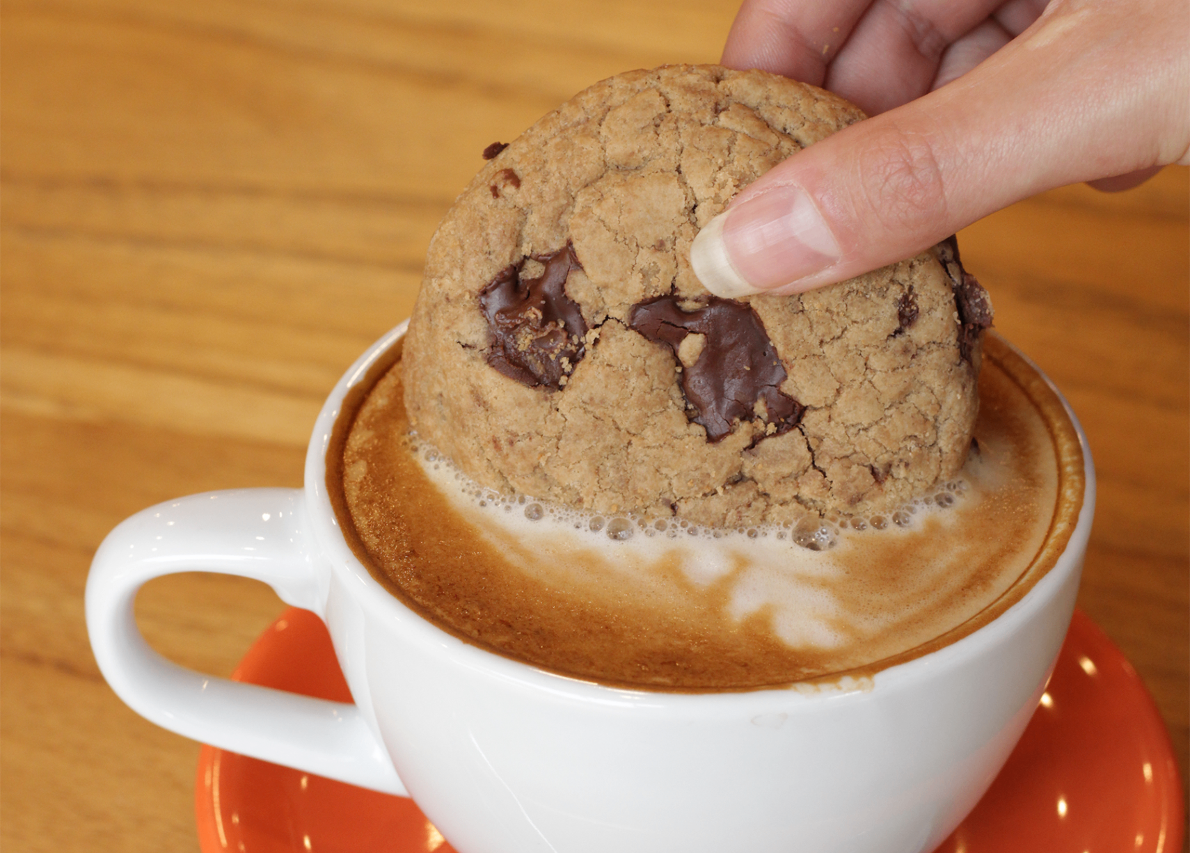 Mão mergulhando cookie de chocolate com pedaços derretidos em xícara branca de cappuccino sobre pires laranja e mesa de madeira do Yarn.