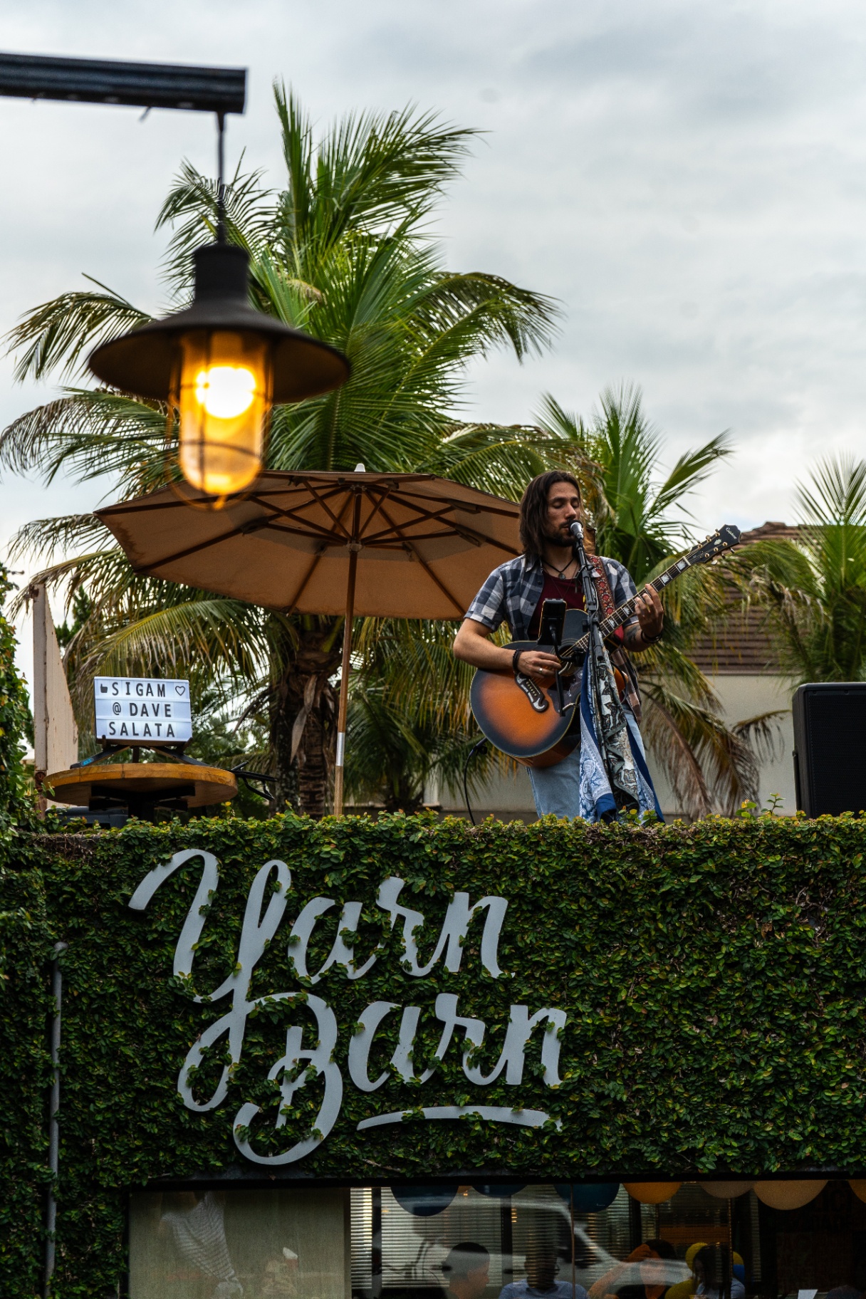 Músico tocando violão e cantando na área externa do Yarn, ao lado do letreiro em folhagem viva, sob luminária acesa e coqueiros ao entardecer.