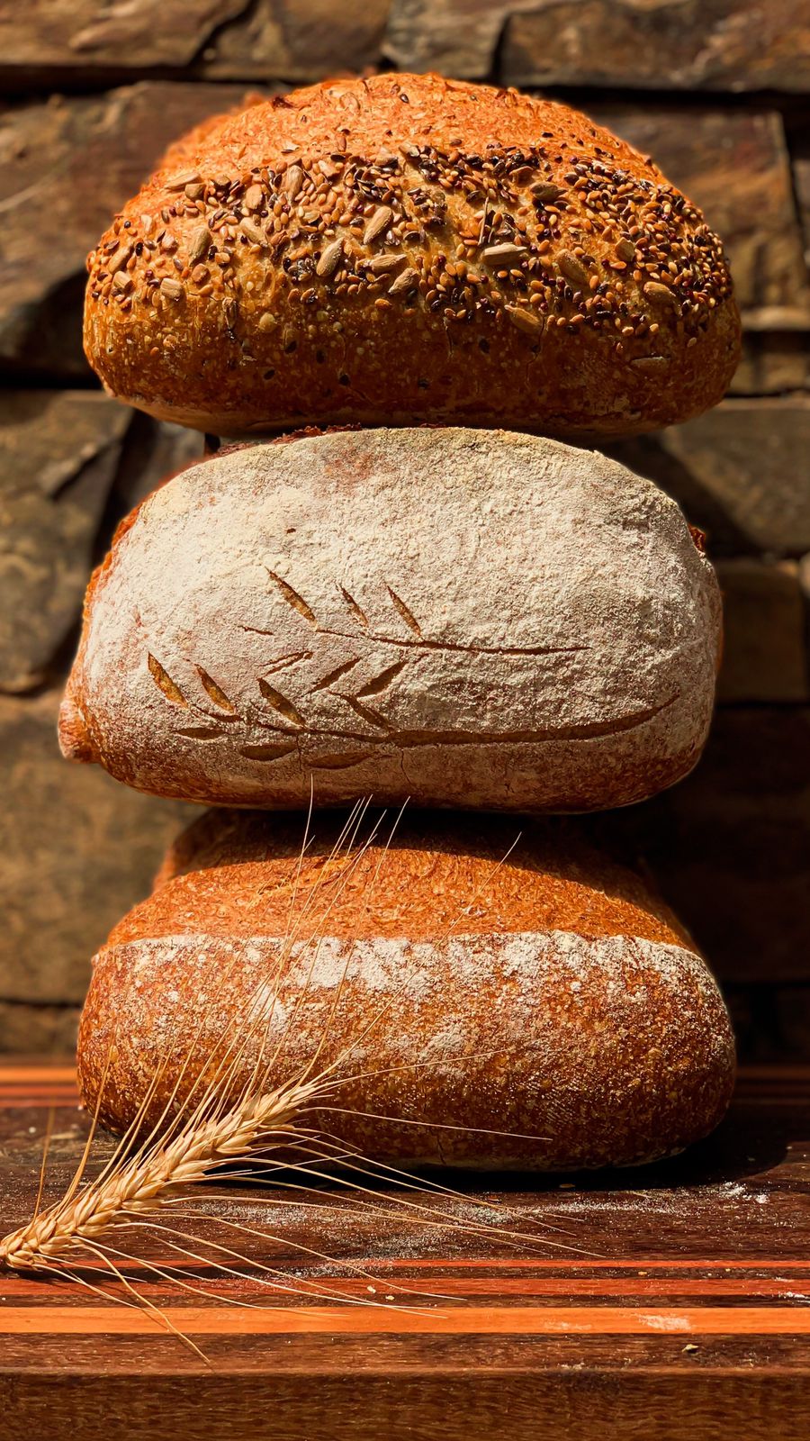 Três pães artesanais de fermentação natural lado a lado, com espigas de trigo, sobre parede de pedra rústica