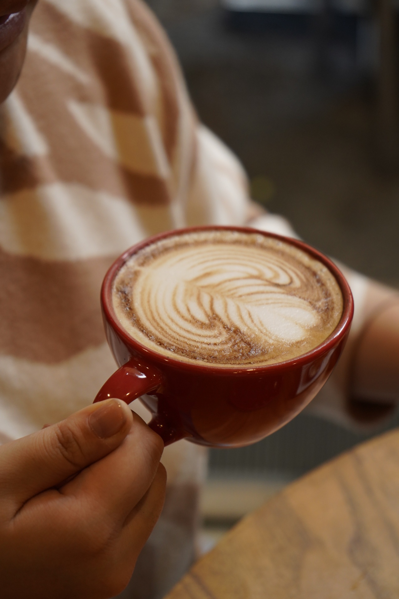 Mão segurando xícara vermelha de café expresso com leite e latte art em folha desenhada na crema, ao fundo pessoa com camisa em xadrez bege e branco no balcão do Yarn.