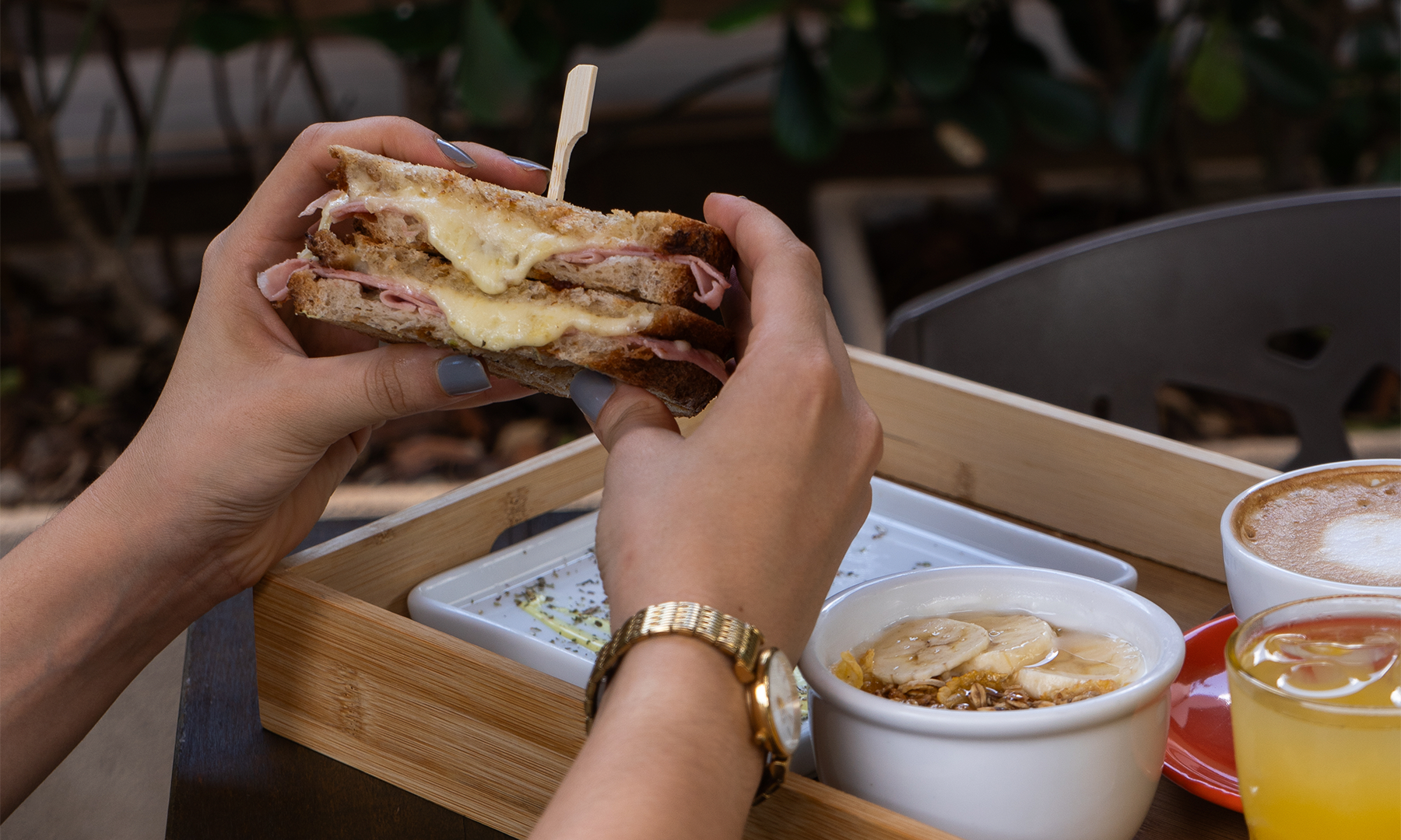 Mãos segurando metade de sanduíche prensado de presunto e queijo derretido do Yarn, com bandeja de madeira ao fundo trazendo iogurte com banana e granola, cappuccino e suco natural.