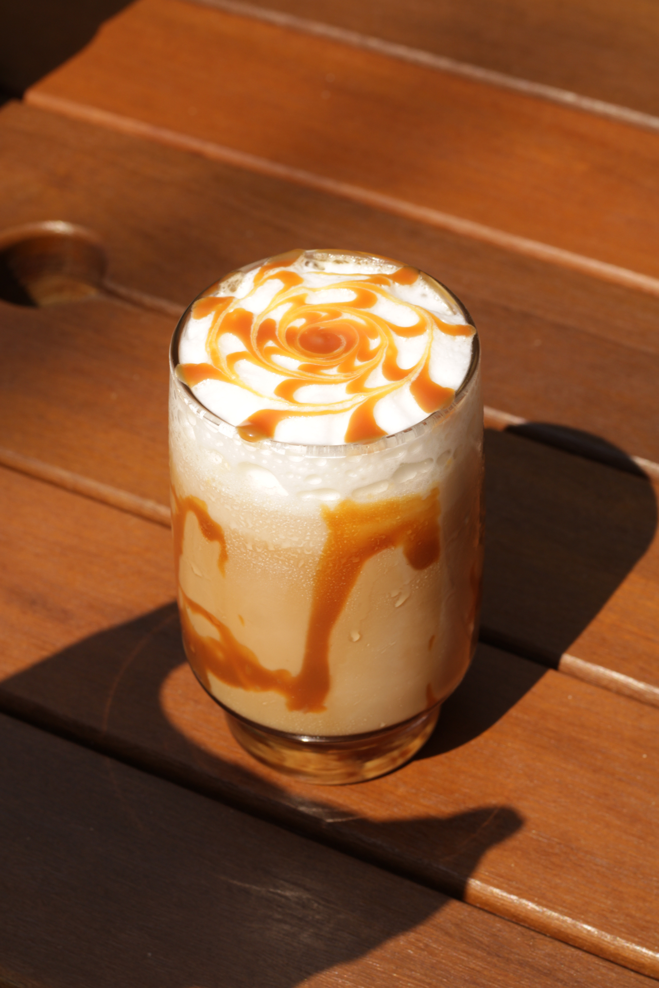 Copo de vidro com café gelado cremoso, espuma branca no topo e desenho em espiral de calda de caramelo, sobre mesa de madeira ripada iluminada pelo sol no Yarn.