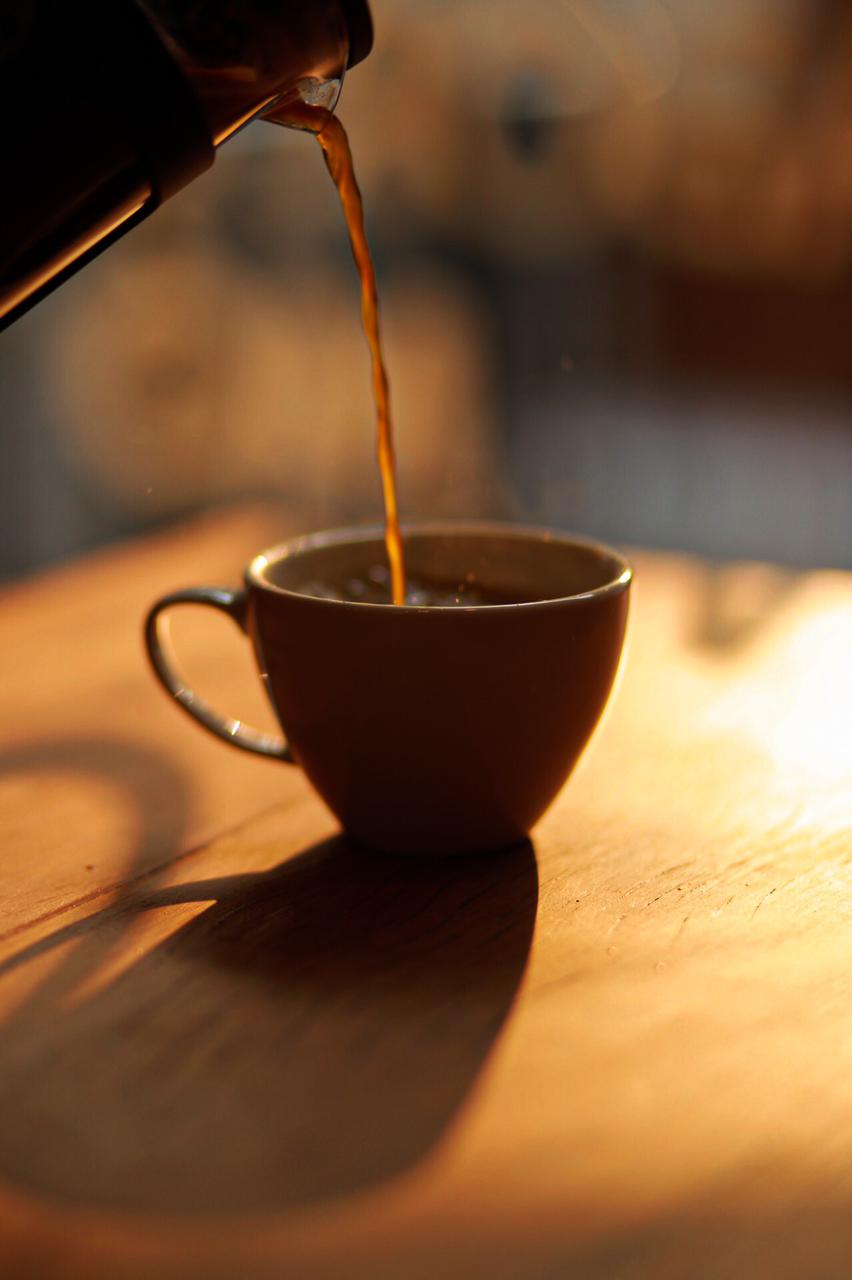 Café sendo despejado em xícara pequena sobre mesa de madeira banhada por luz dourada do fim de tarde.