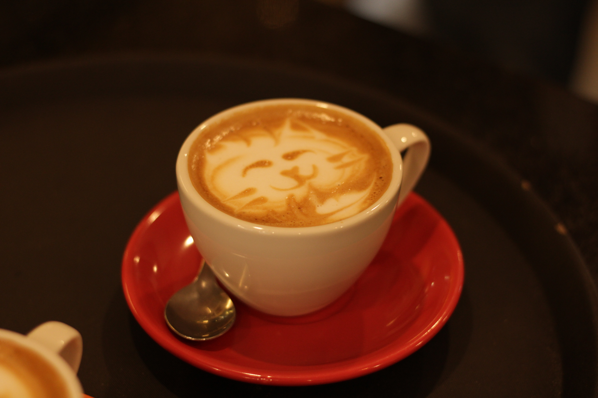 Xícara branca com latte art em forma de gatinho servida em pires vermelho sobre bandeja escura no Yarn.