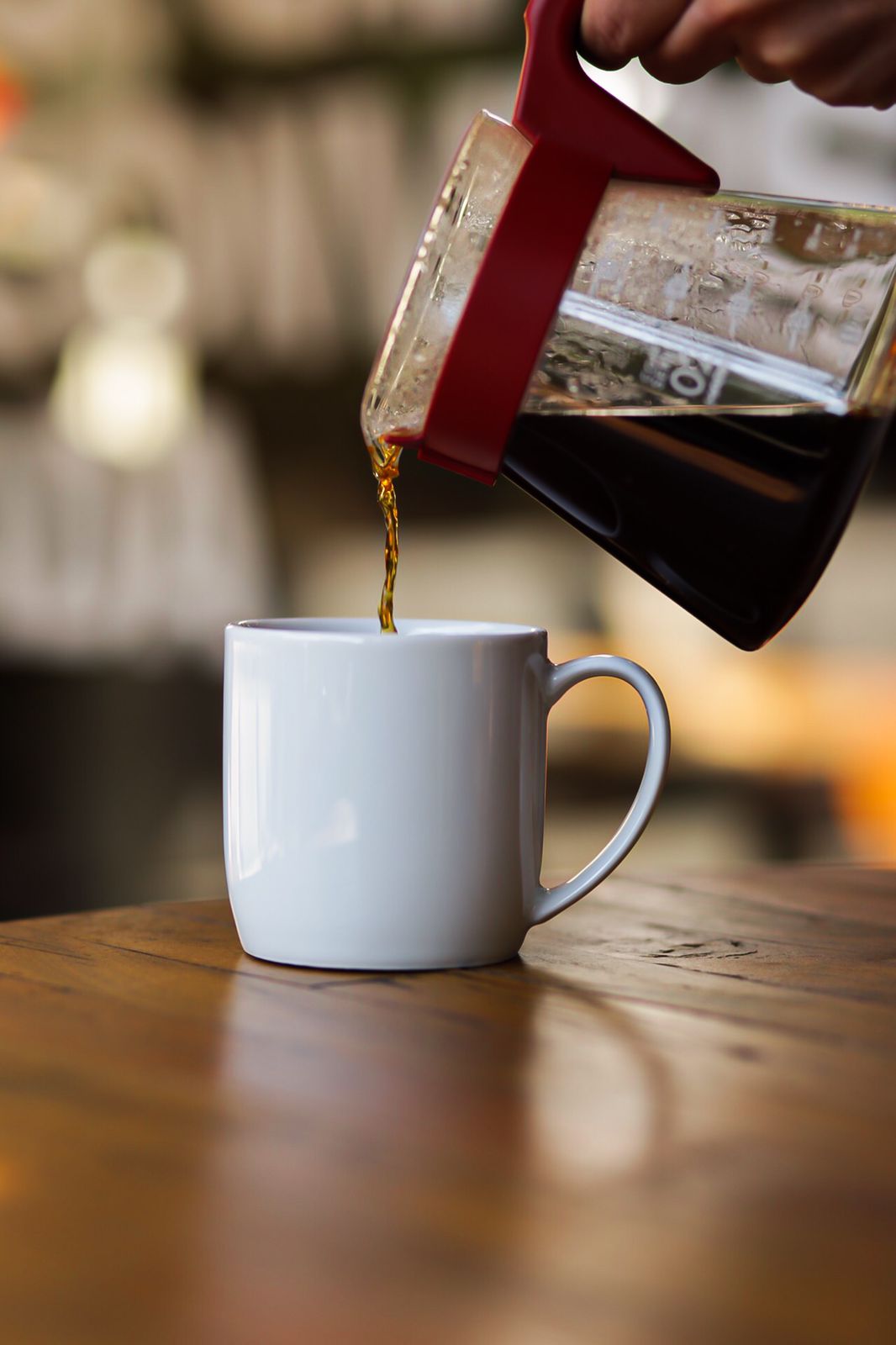 Cafeteira de prensa francesa vermelha servindo café coado em caneca branca sobre mesa de madeira.