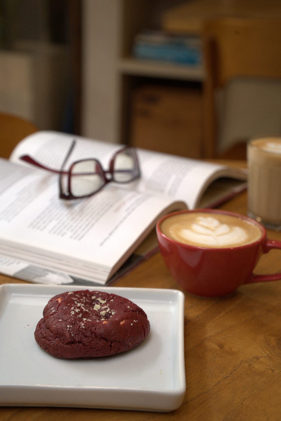 Mesa de madeira do Yarn com cookie red velvet no prato branco, xícara vermelha de cappuccino com latte art, copo de café com leite e livro aberto com óculos de leitura ao lado.