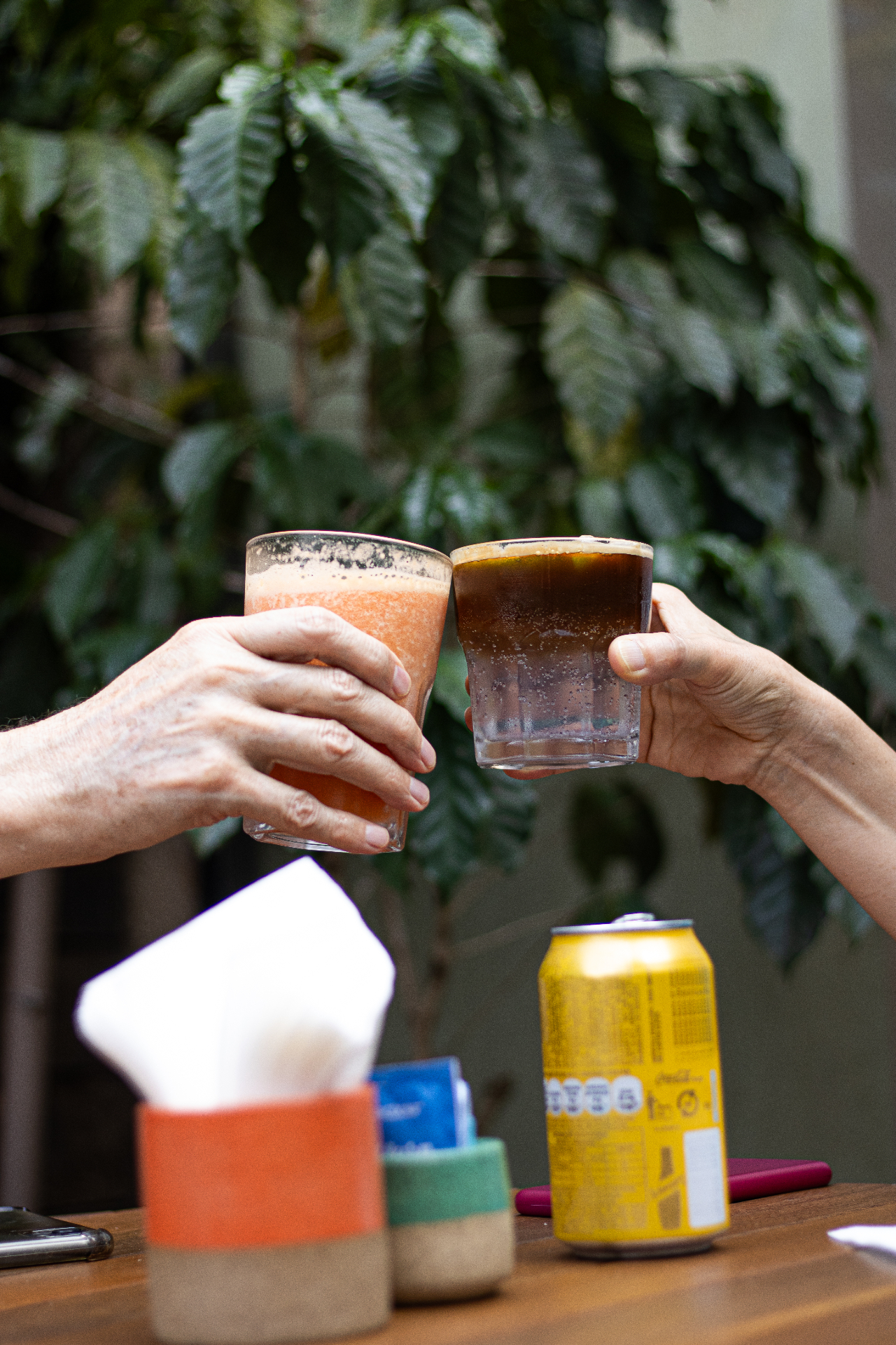 Duas mãos brindando com copos de suco de laranja e café gelado sobre a mesa do Yarn, com folhagem de cafeeiro ao fundo.