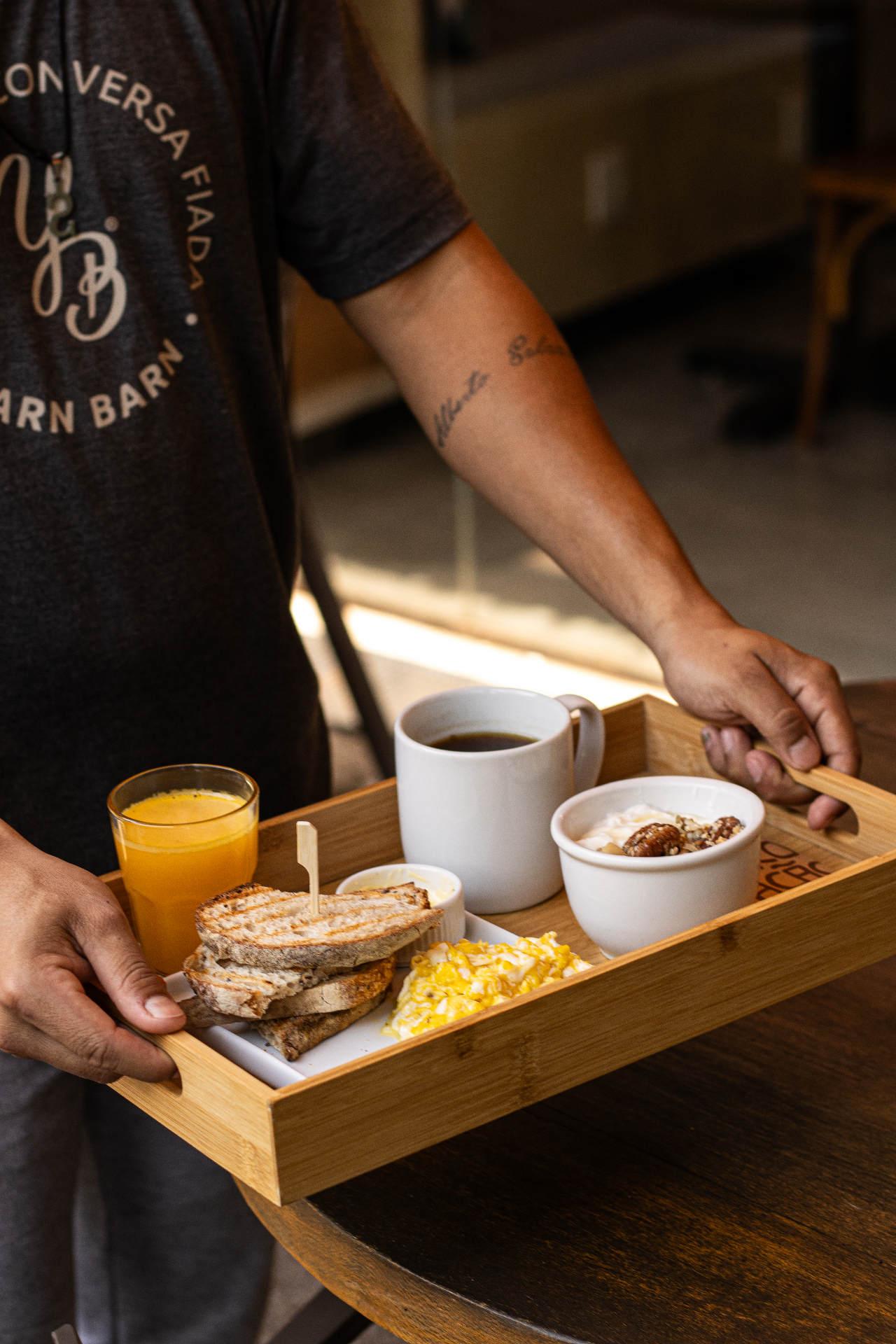 Atendente do Yarn segurando bandeja de bambu com torradas de pão da casa, ovos mexidos, tigelinha de iogurte com granola e nozes, xícara de café preto e copo de suco de laranja.